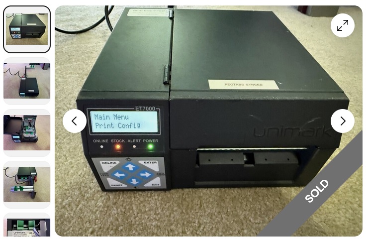 it's a top-front view of a black metal box with a small LCD character display on the front, a membrane keyboard, a spot for a ticket to roll out, the Unimark logo, some status LEDs, and a label that says PECTABS SYNCED. The printer is sitting on carpet, because electronics love sitting on carpet. Static electricity has never harmed any electronics. Thermal printheads are most certainly not known for being highly sensitive to ESD.